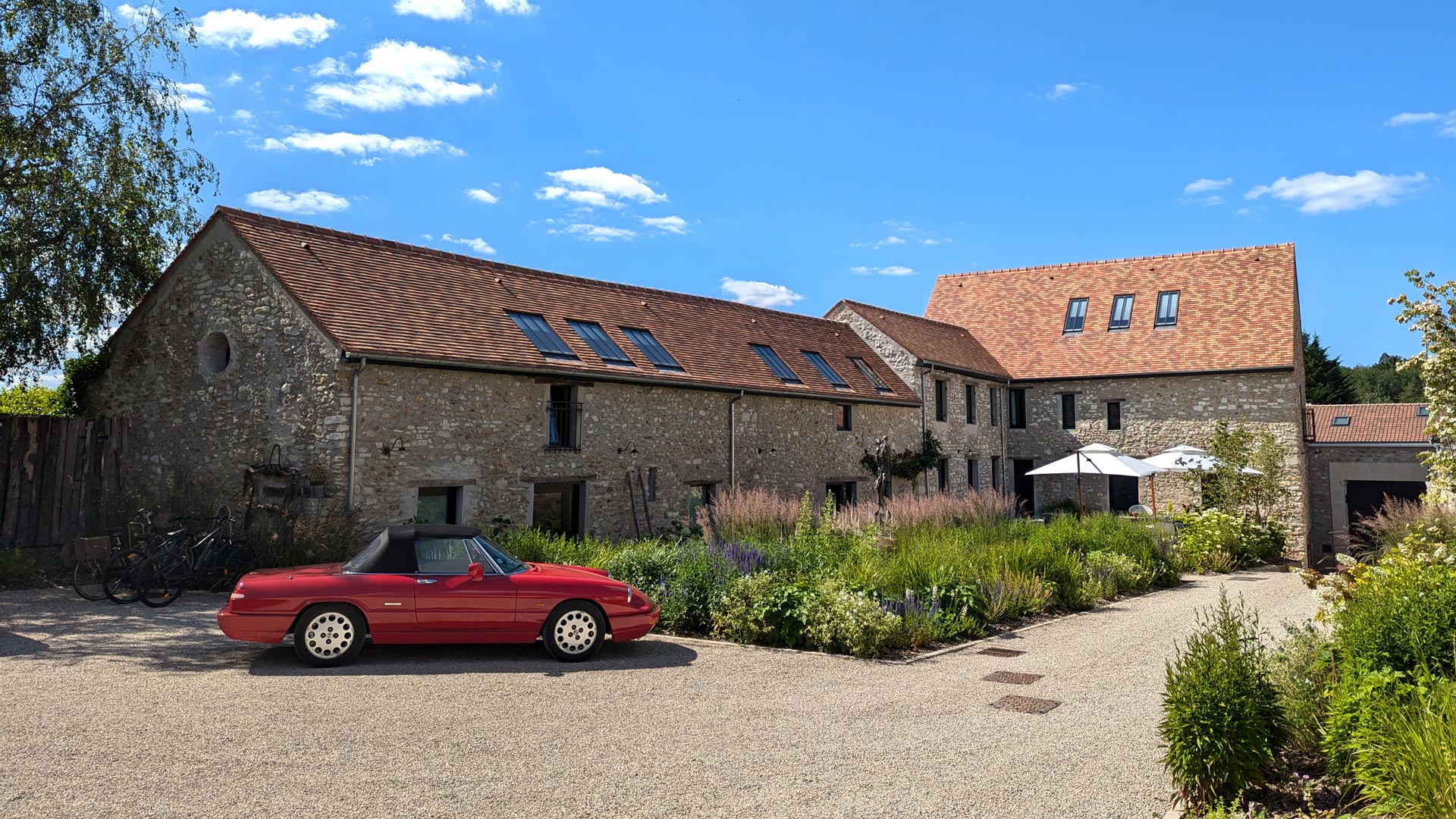 Le bâtiment en pierre rénové avec toit en tuiles rouges de la maison d'hôtes Val Trebbia, entouré d'un jardin fleuri et d'une allée gravillonnée, avec une Alpha Romeo Spider rouge et décapotable stationnée à l'avant sous un ciel bleu dégagé.