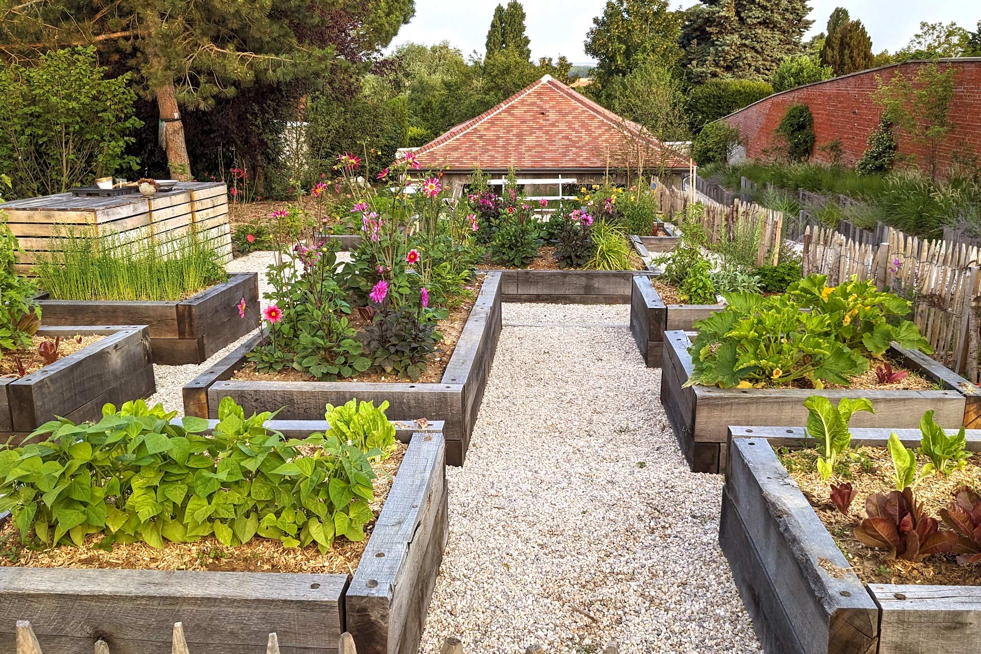 Potager de la maison d'hôtes Val Trebbia, organisé en carrés surélevés en bois, avec des allées gravillonnées, des légumes et des fleurs colorées, entouré de végétation luxuriante et d'une clôture en bois.