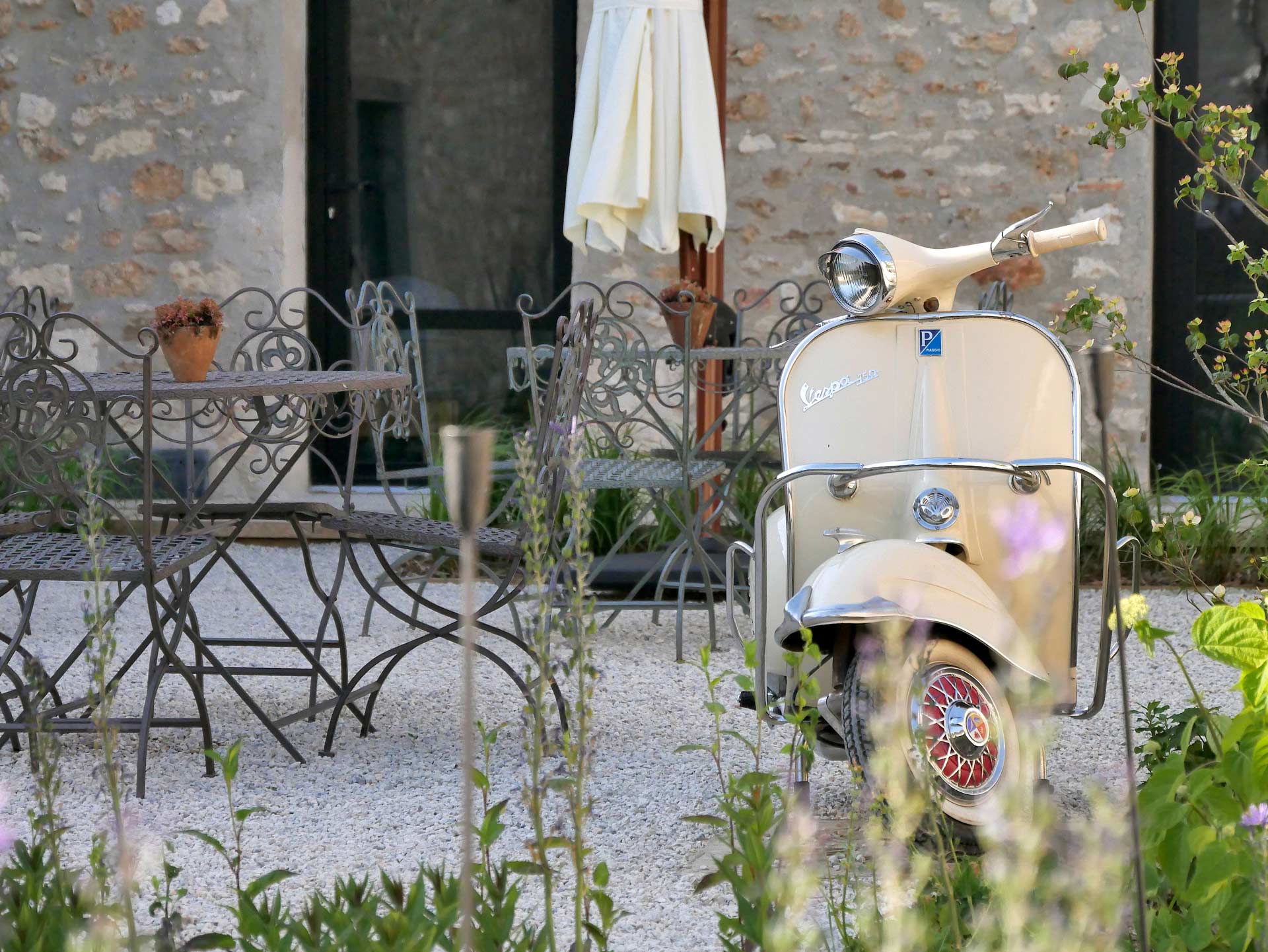 Scooter Vespa crème utilisé comme élément décoratif, placé sur une terrasse en gravier, près d'une table et de chaises en fer forgé, dans le jardin de la maison d'hôtes Val Trebbia