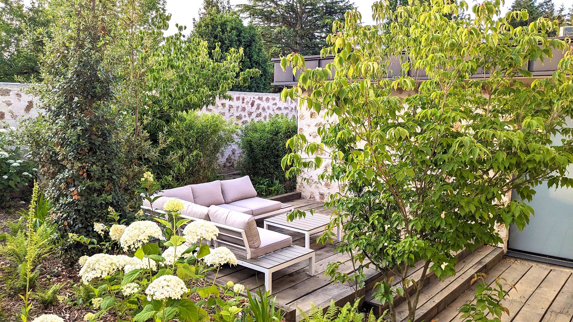 Coin détente extérieur avec un canapé d'angle beige sur une terrasse en bois, entouré de verdure luxuriante et de fleurs blanches, dans un jardin bordé d'un mur en pierre, derrière le spa de la maison d'hôtes Val Trebbia